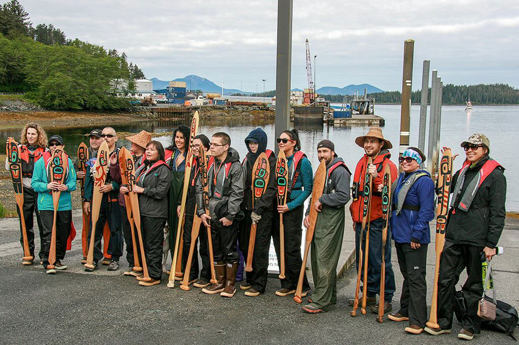 Community members standing on a dock holding decorated canoe paddles